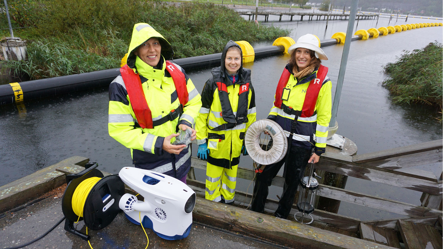 tre personer i regntøy, bak undervannsdrone.